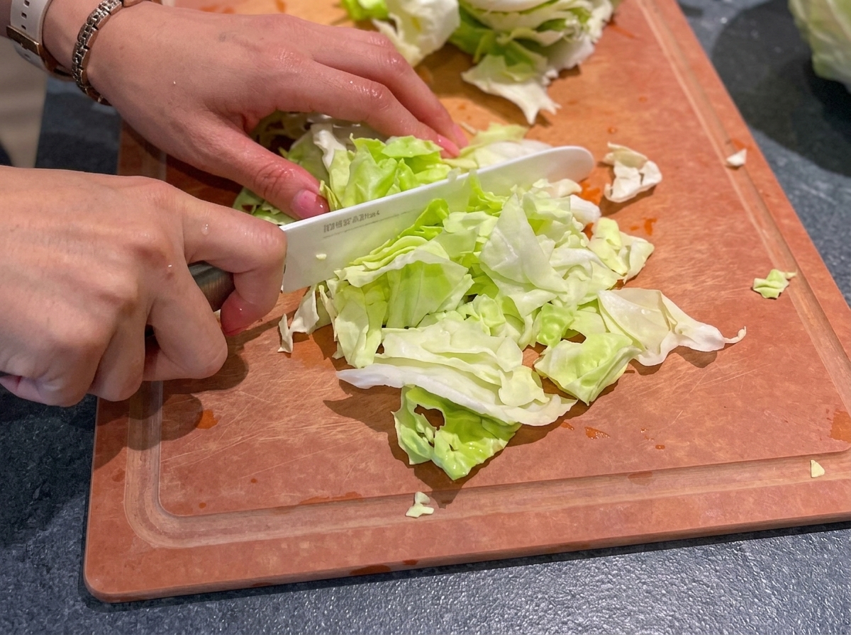 Mains utilisant un couteau en céramique blanc pour couper du chou vert frais sur une planche à découper en bois.