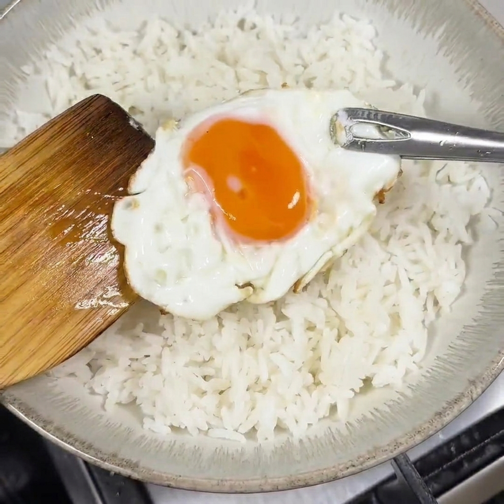 Une spatule en bois déposant un œuf au plat avec un jaune coulant sur un bol de riz blanc.