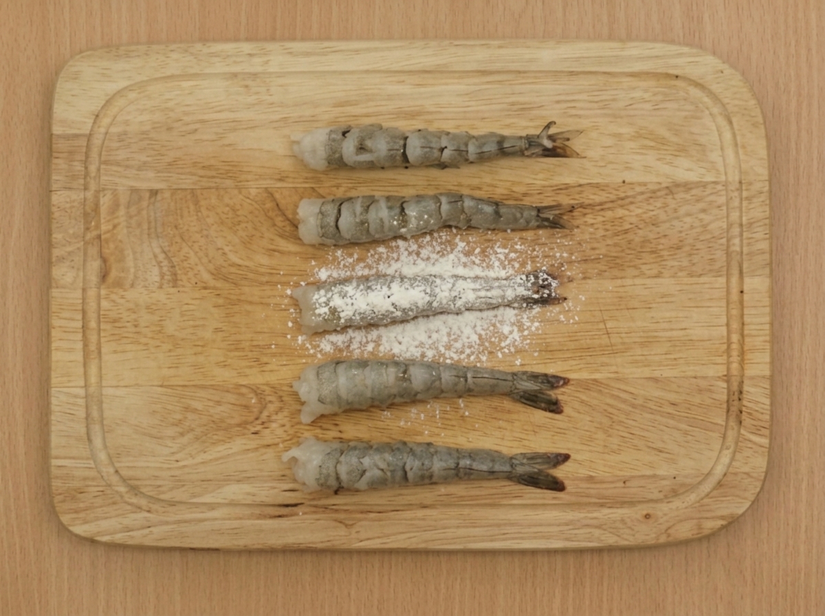 Un tamis saupoudrant de la farine sur plusieurs crevettes préparées et disposées sur une planche en bois.