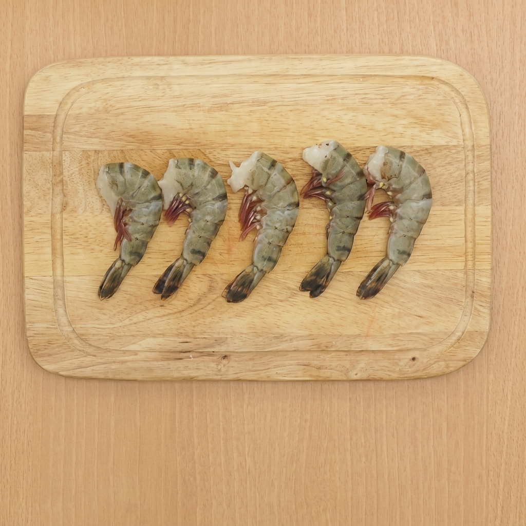 Cinq crevettes nettoyées sur une planche à découper en bois, avec leurs queues intactes.