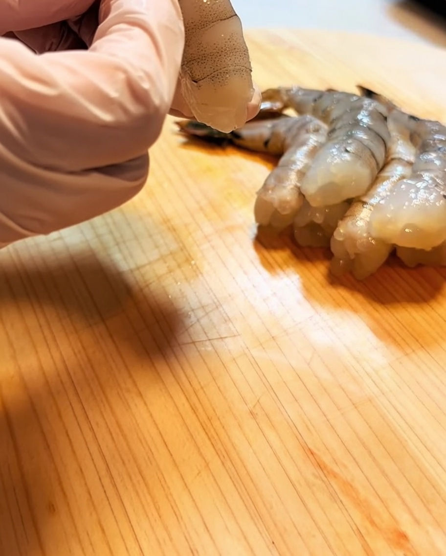 Une main gantée de rose tenant une crevette nettoyée, déveinée et préparée sur une surface en bois.
