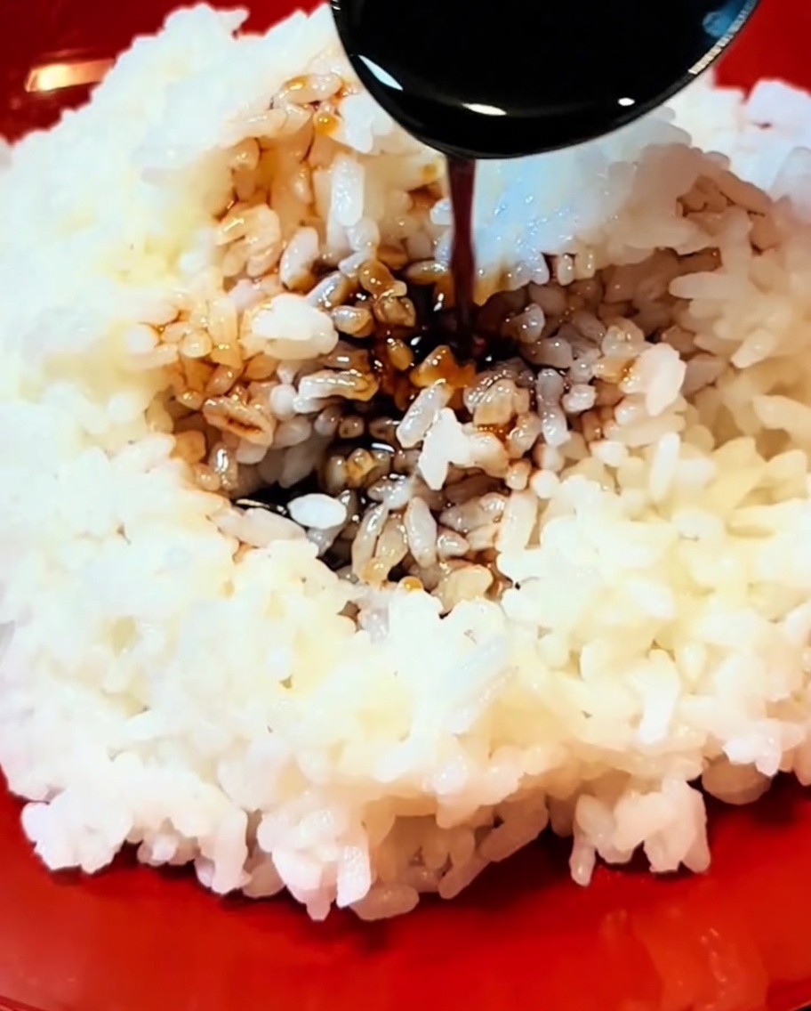 Gros plan sur une sauce épaisse sombre versée dune cuillère sur une portion de riz blanc moelleux dans un bol rouge.