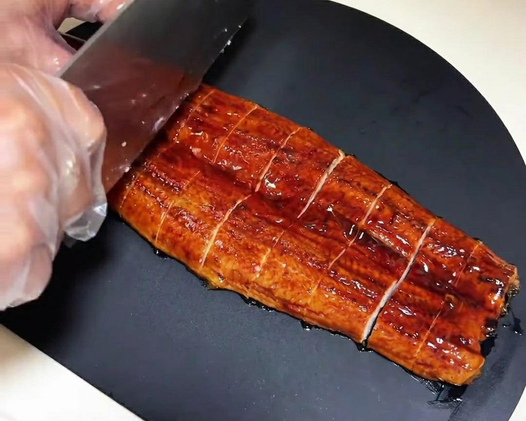 Un filet danguille pré-grillé étant coupé en morceaux rectangulaires uniformes sur une planche à découper foncée.