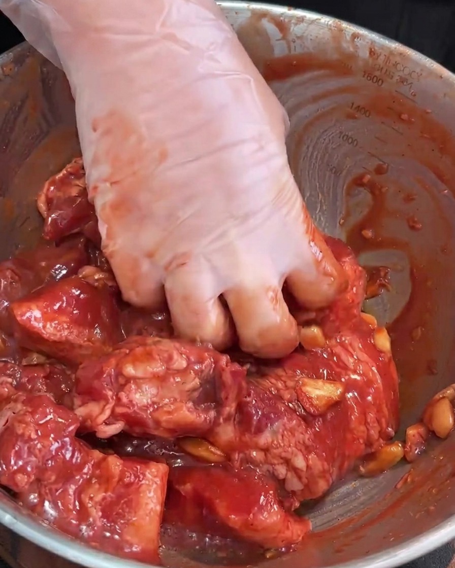 Des mains gantées massant la marinade dans les lanières de porc crues dans un saladier en inox.