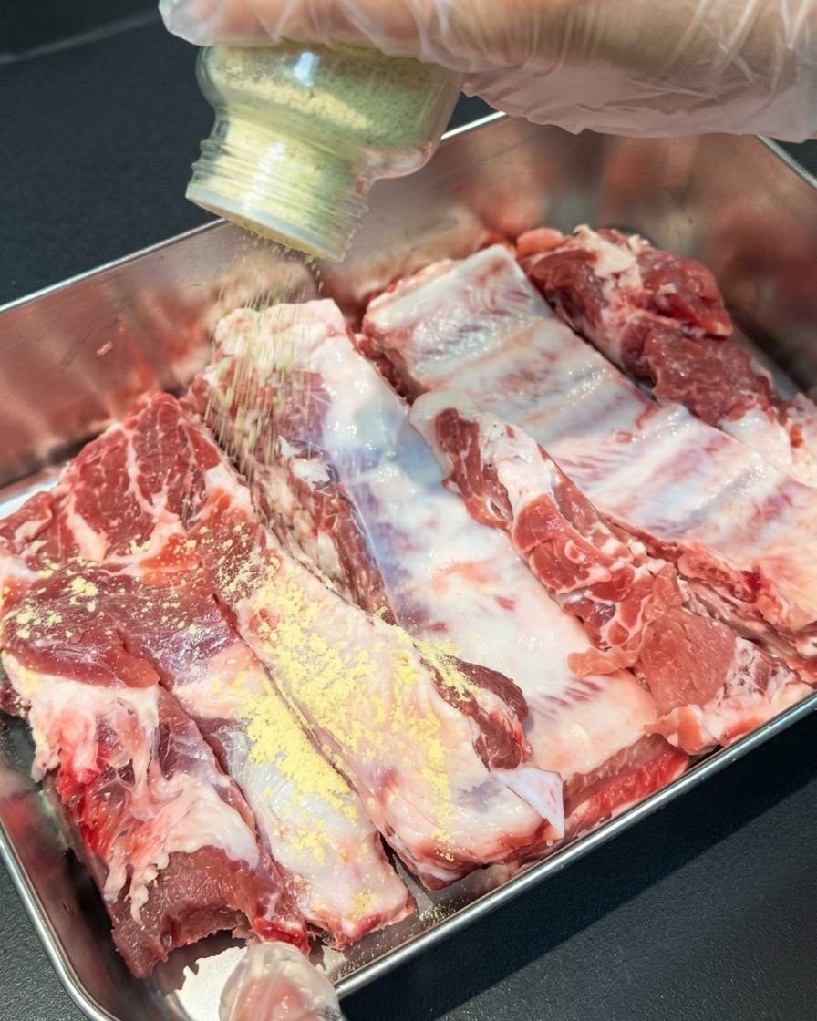 Une main saupoudrant de lail et du gingembre en poudre à partir dun flacon en verre sur des travers de porc crus dans un plateau en métal.