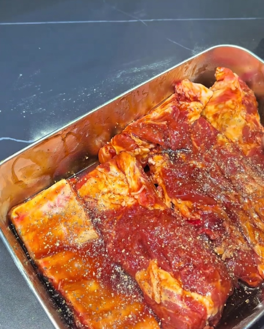Gros plan de travers de porc crus entièrement enrobés dune marinade sombre et saupoudrés de flocons de poivre noir concassé.