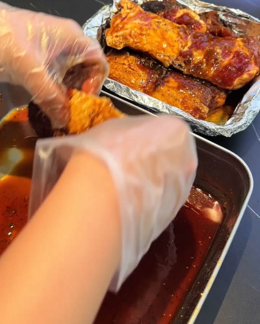 Mains gantées transférant des travers de porc marinés dans un récipient tapissé de papier daluminium pour la cuisson à la vapeur.
