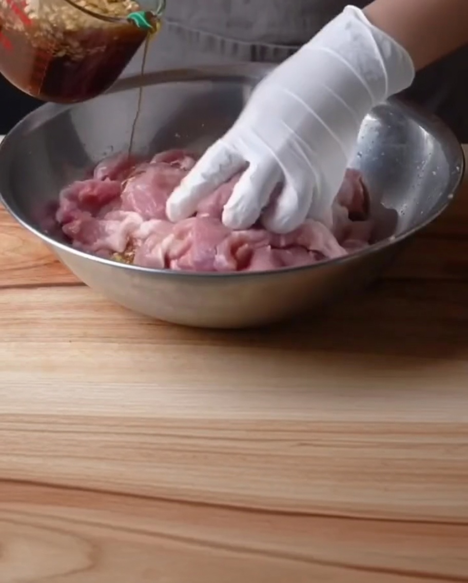 Une main gantée versant une marinade sombre et assaisonnée sur des travers de porc dans un grand bol en métal.