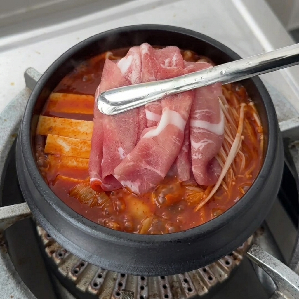 Des tranches de porc frais disposées sur le dessus des ingrédients du Kimchi Jjigae qui mijotent.