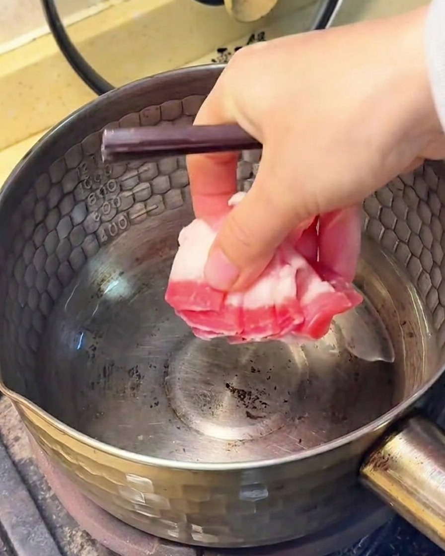 Tranches de poitrine de porc frisant dans une casserole en inox, la graisse fondue est visible au fond.
