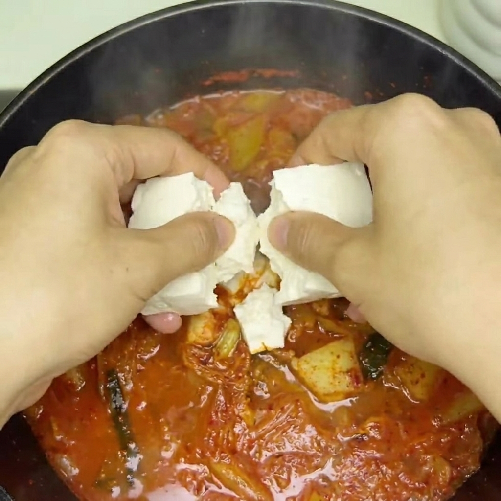 Des cubes de tofu frais délicatement placés dans la soupe au kimchi bouillonnante.