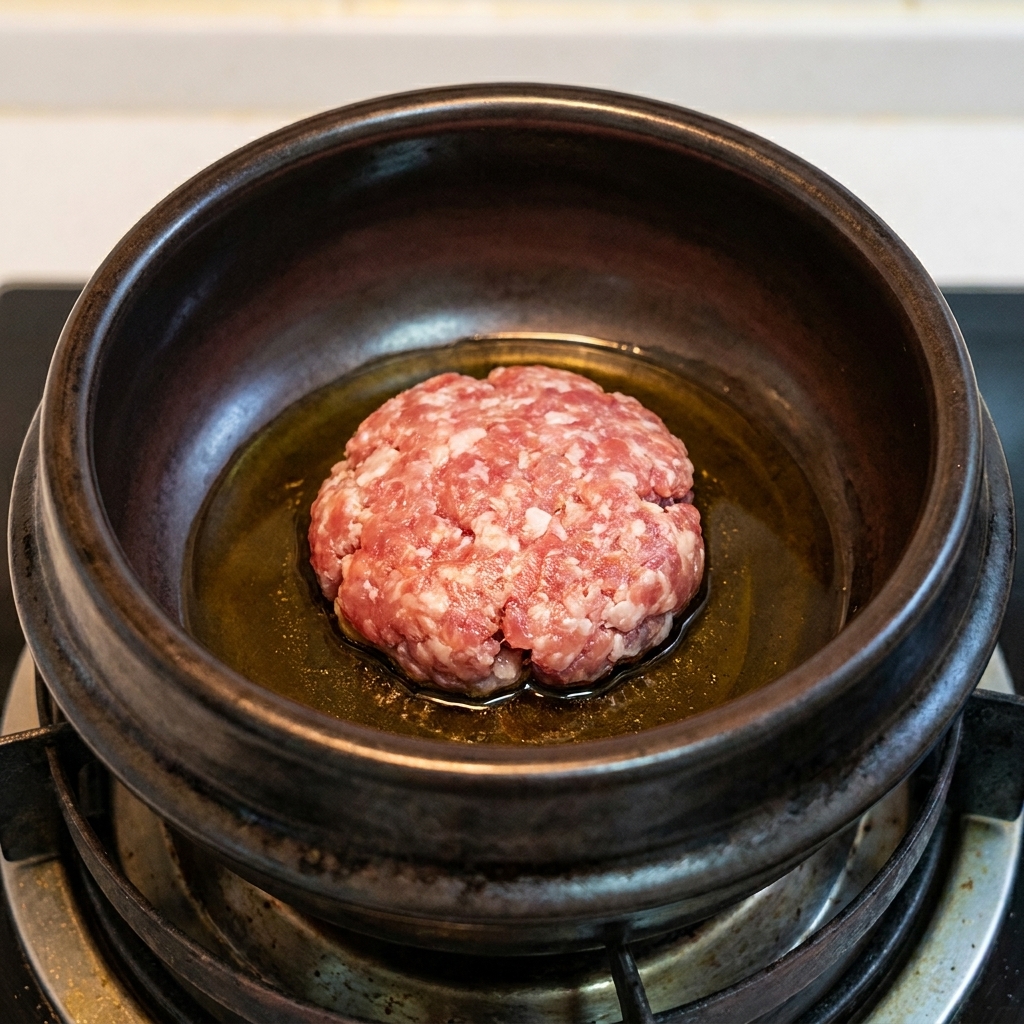 Une boule de porc haché brunissant dans lhuile de sésame chaude à lintérieur dun pot en terre cuite noir.