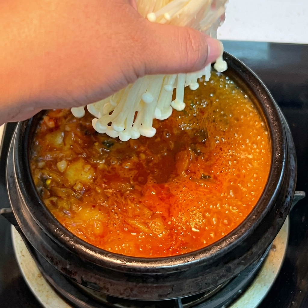 Une main plaçant une botte de champignons enoki frais dans le ragoût de kimchi rouge bouillonnant.