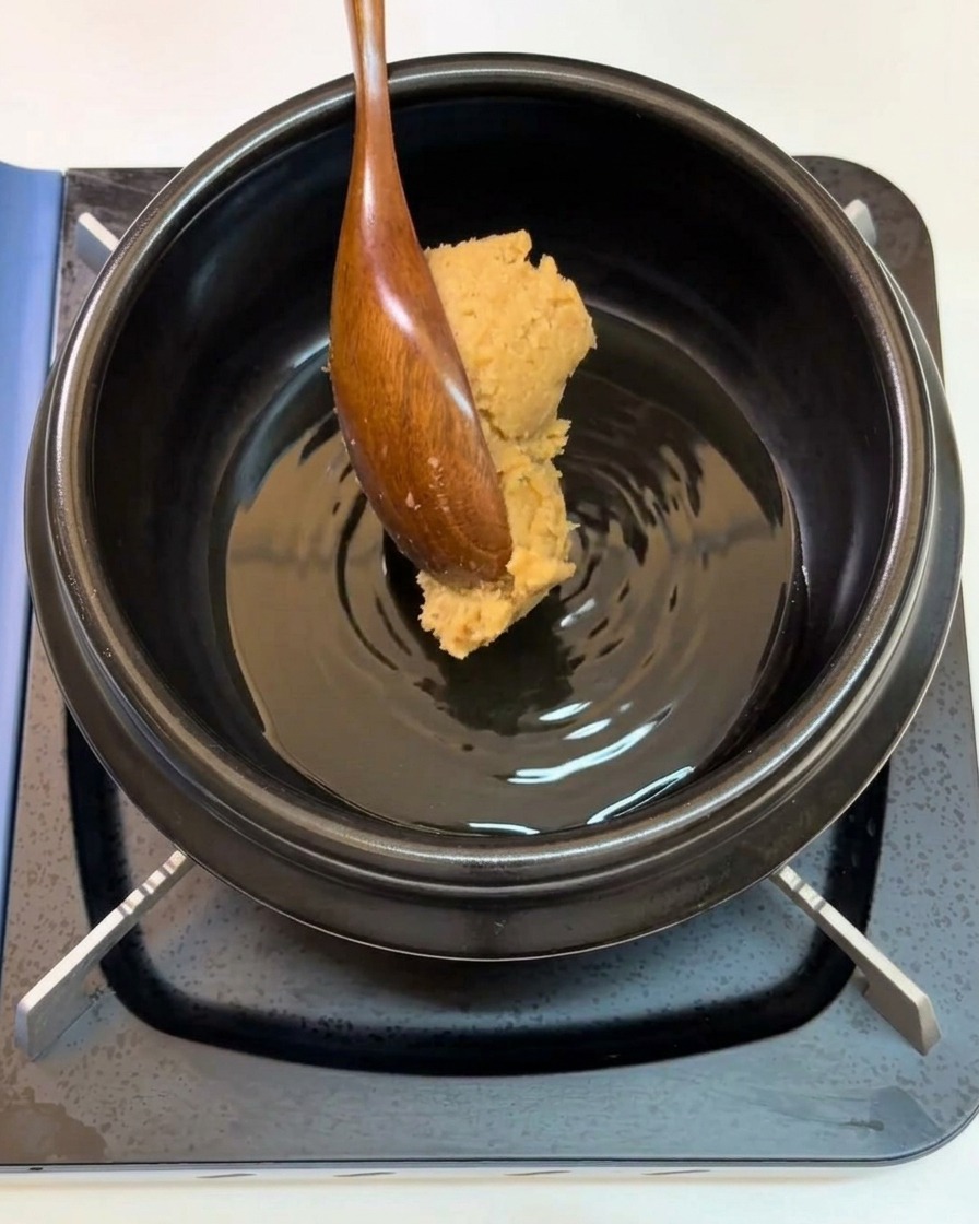 Une cuillère en bois ajoutant une noisette de pâte de soja dans un pot.
