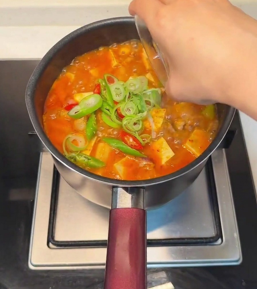 Une main saupoudrant des oignons nouveaux émincés et des piments frais rouges et verts sur le ragoût de tofu fini.