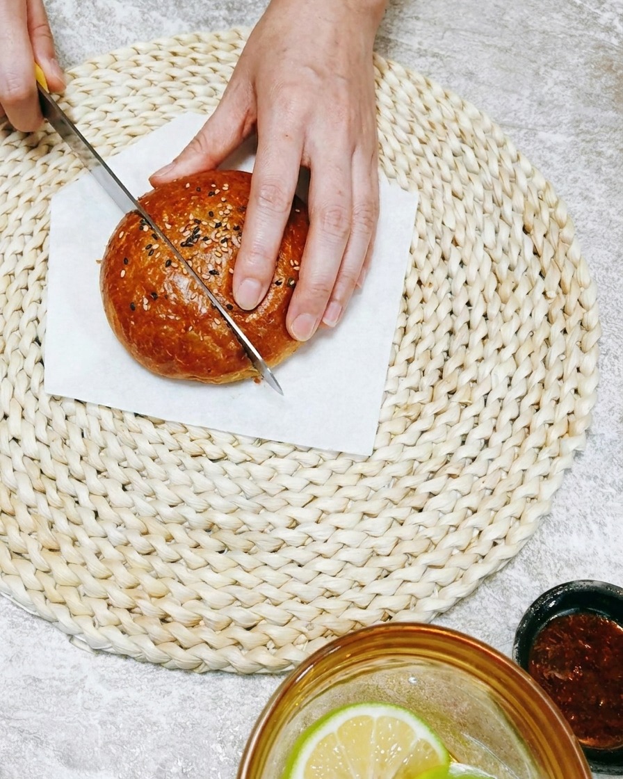 Gros plan sur une main utilisant un couteau pour trancher un pain à burger sur un tapis tressé.