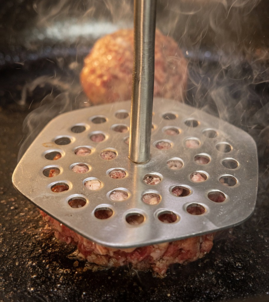 Une presse à viande en métal utilisée pour écraser les boules de bœuf en steaks fins dans la poêle chaude.