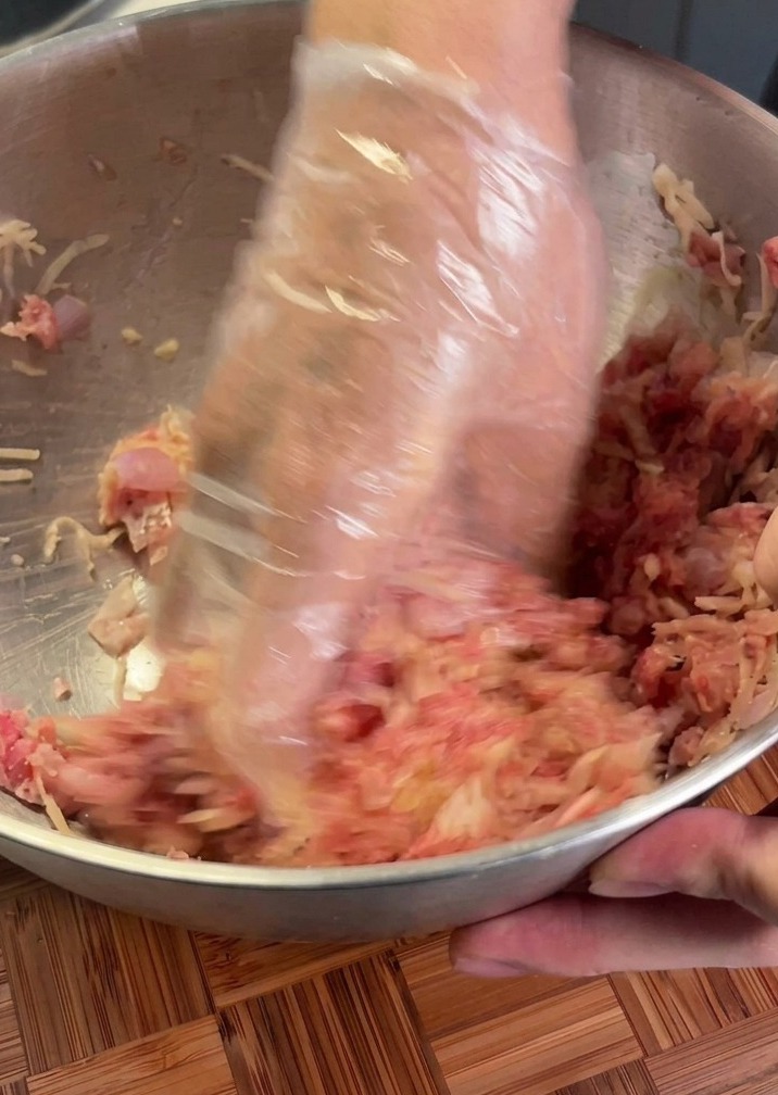 Mains avec un gant en plastique mélangeant le bœuf cru et la pomme de terre râpée dans un bol en métal.