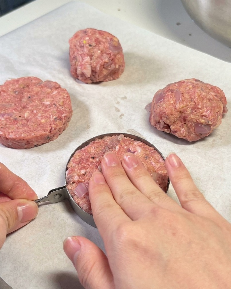 Mains utilisant un cercle de dressage en métal pour façonner un steak haché sur du papier sulfurisé.