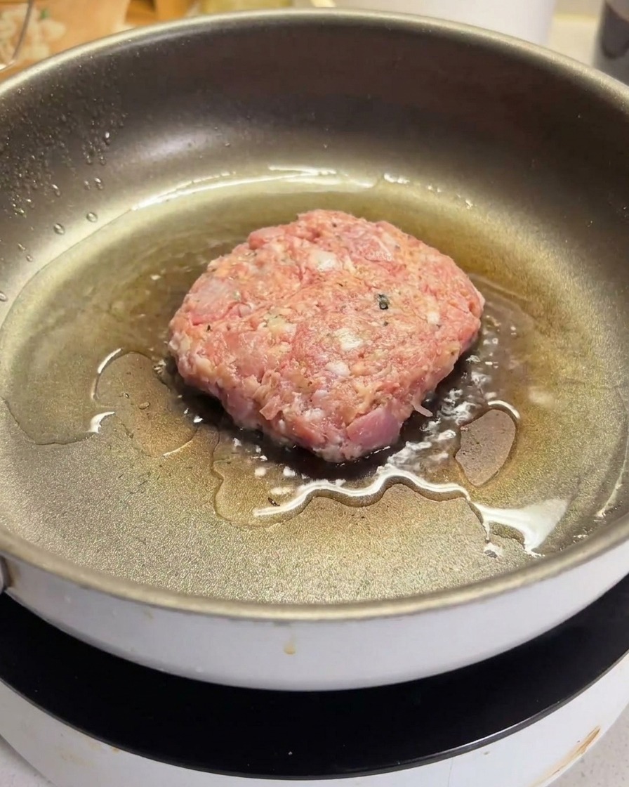 Un steak de bœuf haché cru qui grésille dans une poêle antiadhésive chaude et huilée.