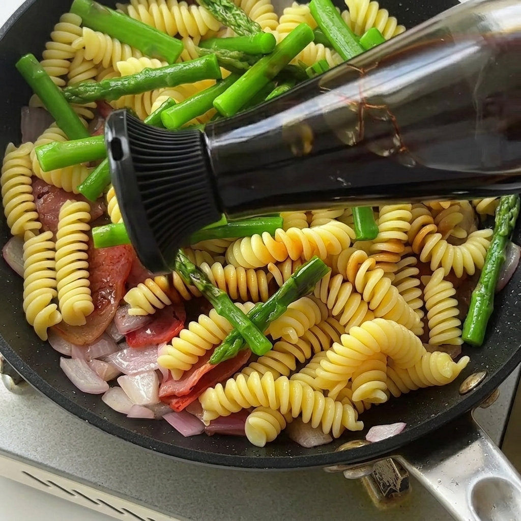 Gros plan sur une poêle contenant les pâtes, les asperges et le bacon, assaisonnés avec une sauce riche et sombre.