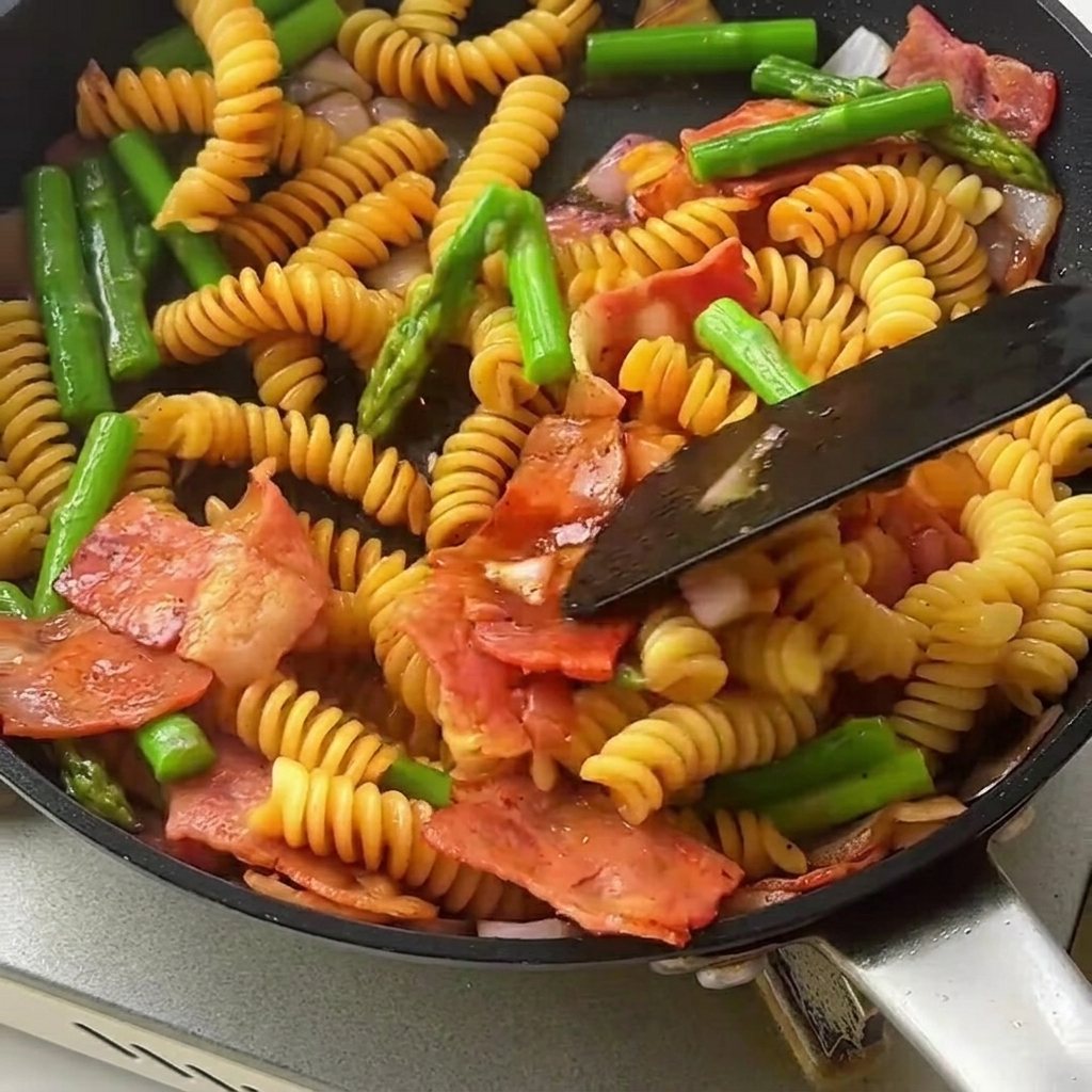 Une spatule mélange les pâtes nappées, les asperges croquantes et le bacon dans une grande poêle.