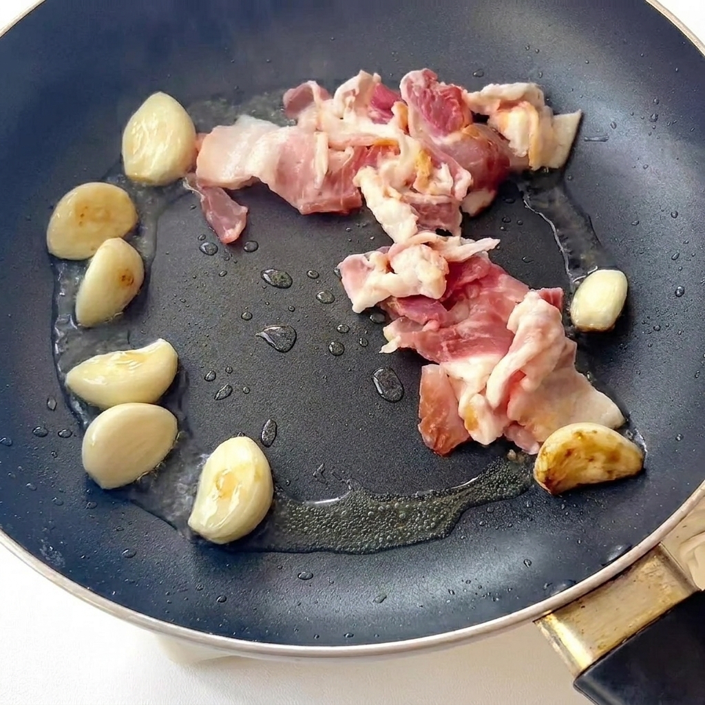 Bacon en train de cuire dans une poêle avec des gousses dail dorées.