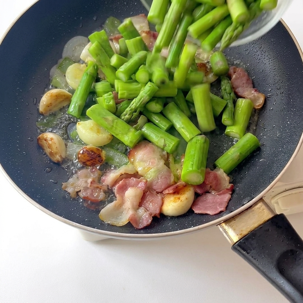 Morceaux dasperges vert vif fraîchement blanchis versés dans une poêle avec de lail et du bacon.