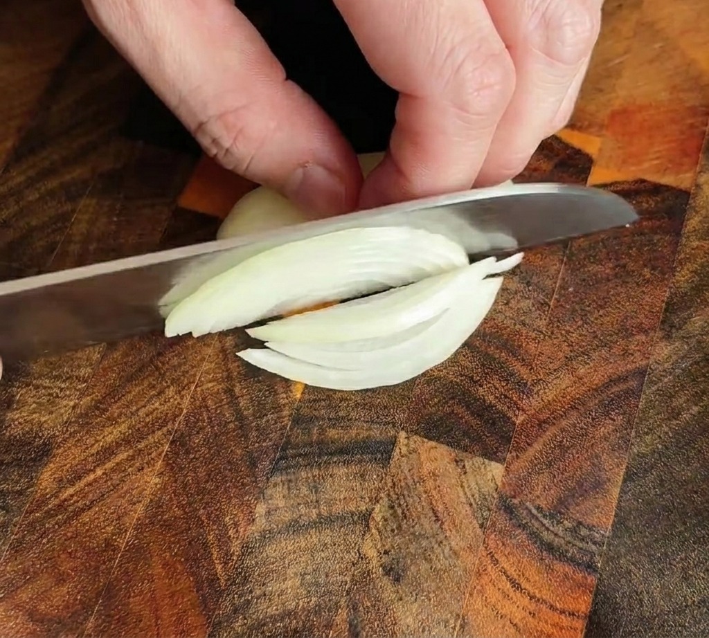 Couteau de chef coupant un oignon en fines lamelles sur une planche en bois.