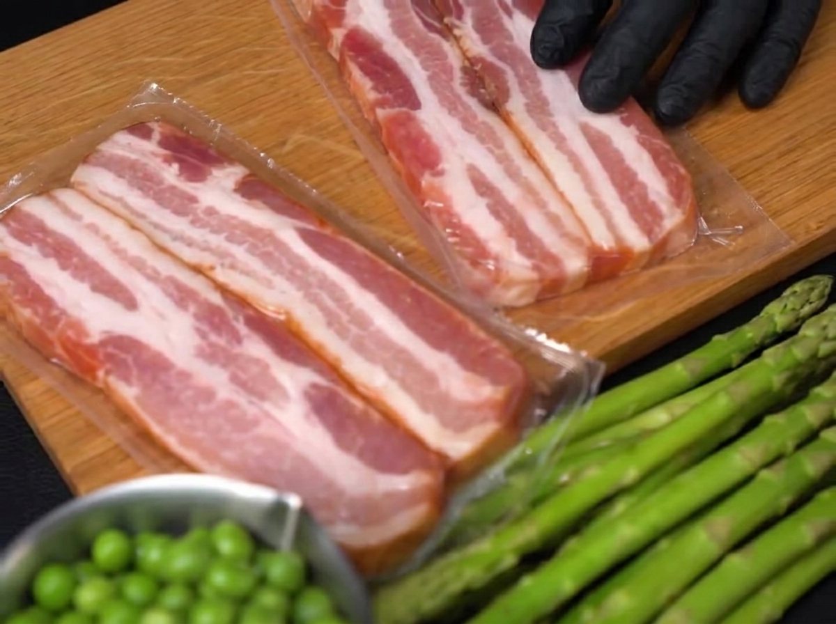 Gros plan sur une main gantée utilisant un couteau pour couper des tranches de bacon sur une planche en bois.