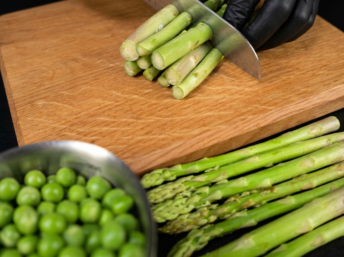 Une main de chef coupant des asperges fraîches en tronçons égaux sur une planche en bois.