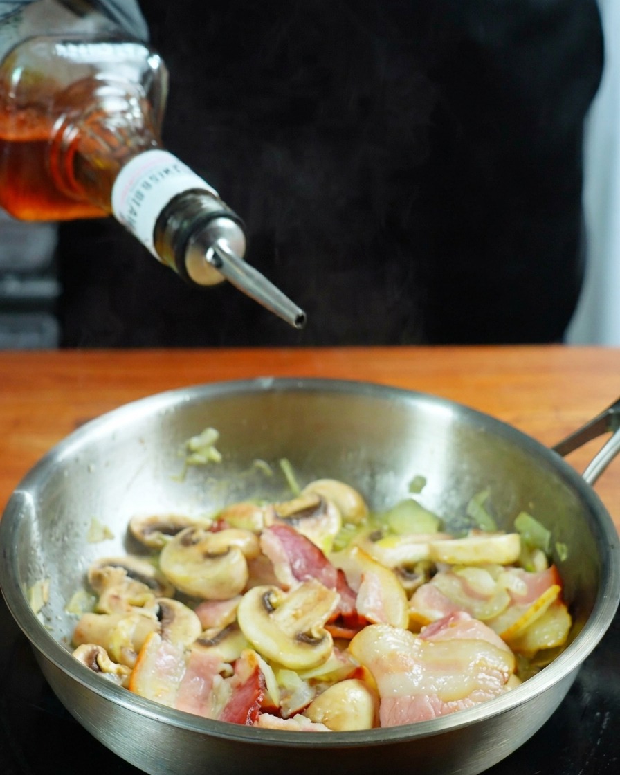 Whisky versé depuis une bouteille dans une poêle contenant le bacon et les champignons.