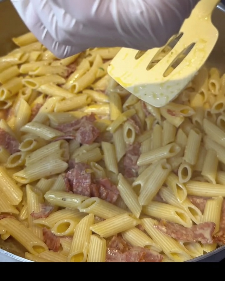 Mains gantées saupoudrant lassaisonnement sur une poêle de pâtes au bacon crémeuses.