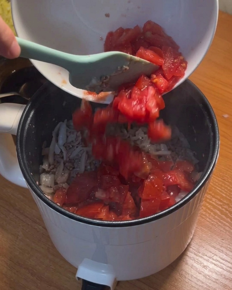 Des tomates en dés versées depuis un bol blanc dans la casserole contenant le bœuf et les oignons sautés.