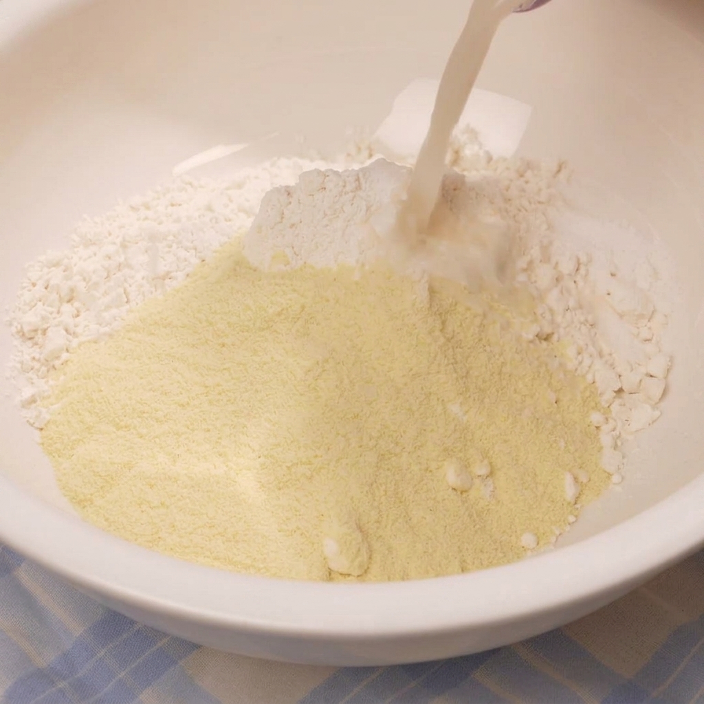 Verser un liquide laiteux contenant la levure dans un bol mélangeur blanc rempli dun monticule de farine et dingrédients secs.