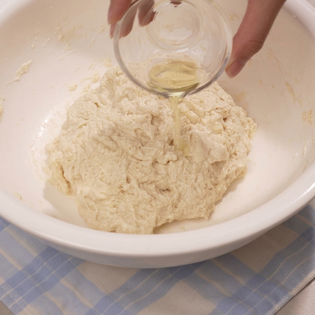 Une main versant de lhuile dun petit bol en verre sur une pâte rugueuse dans un grand bol.