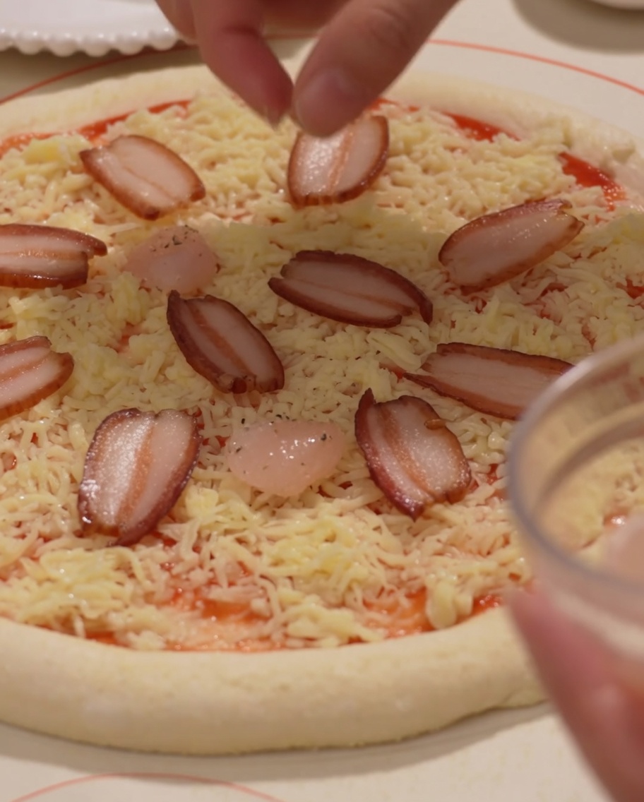 Des tranches de jambon et des crevettes étant soigneusement placées sur une base de pizza garnie de fromage.