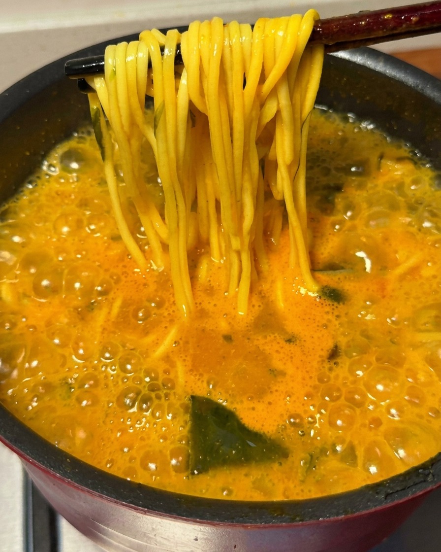 Des baguettes soulevant une botte de nouilles ramen cuites hors du bouillon épicé bouillonnant.