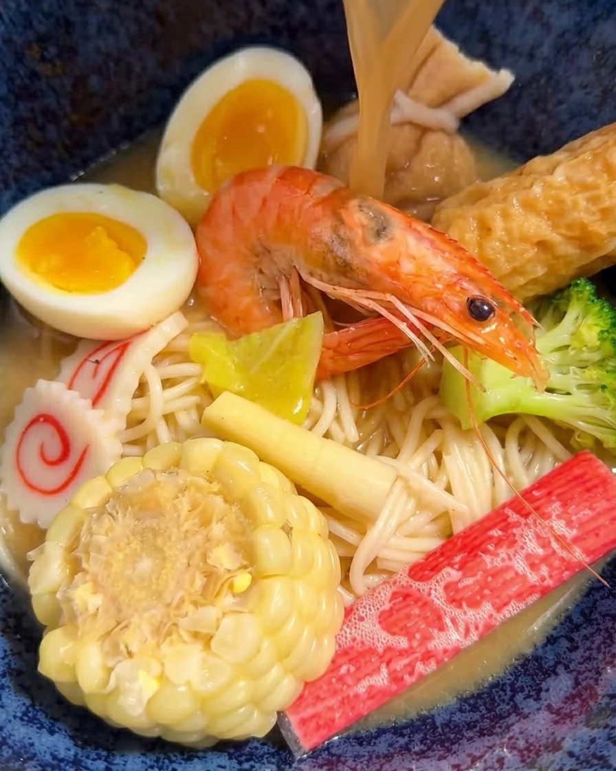 Bouillon miso chaud versé dune louche dans un bol de ramen rempli de diverses viandes et légumes.