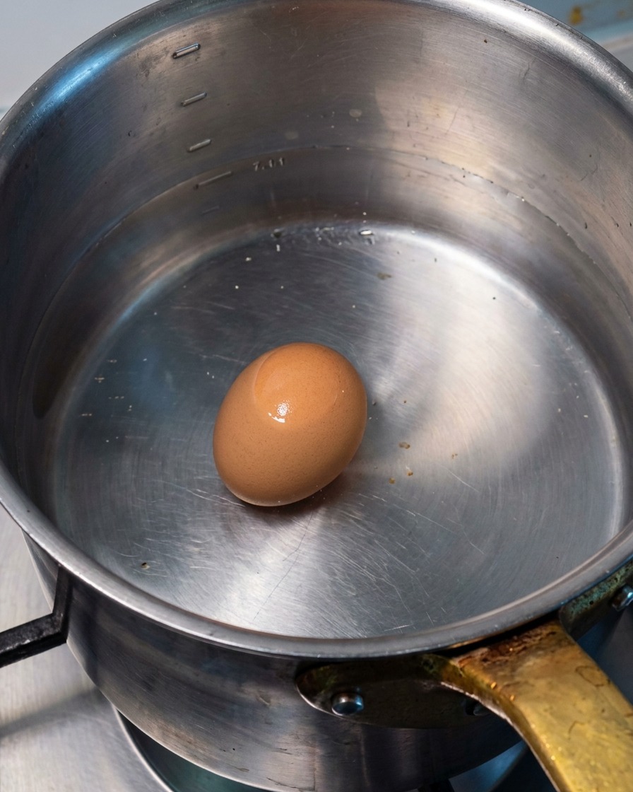 Un œuf entier marron reposant au fond dune casserole en métal remplie deau chaude.