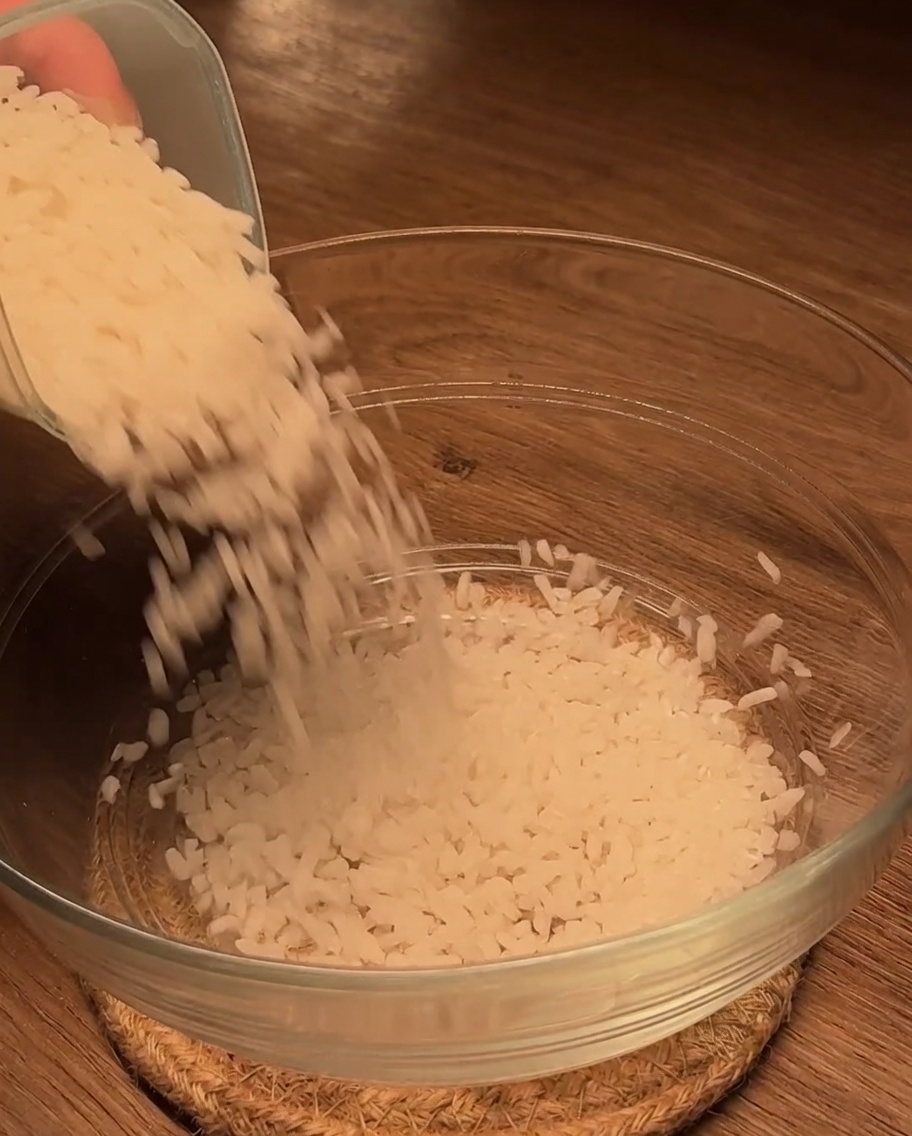 Du riz blanc cru est versé dans un bol en verre transparent sur une surface en bois.