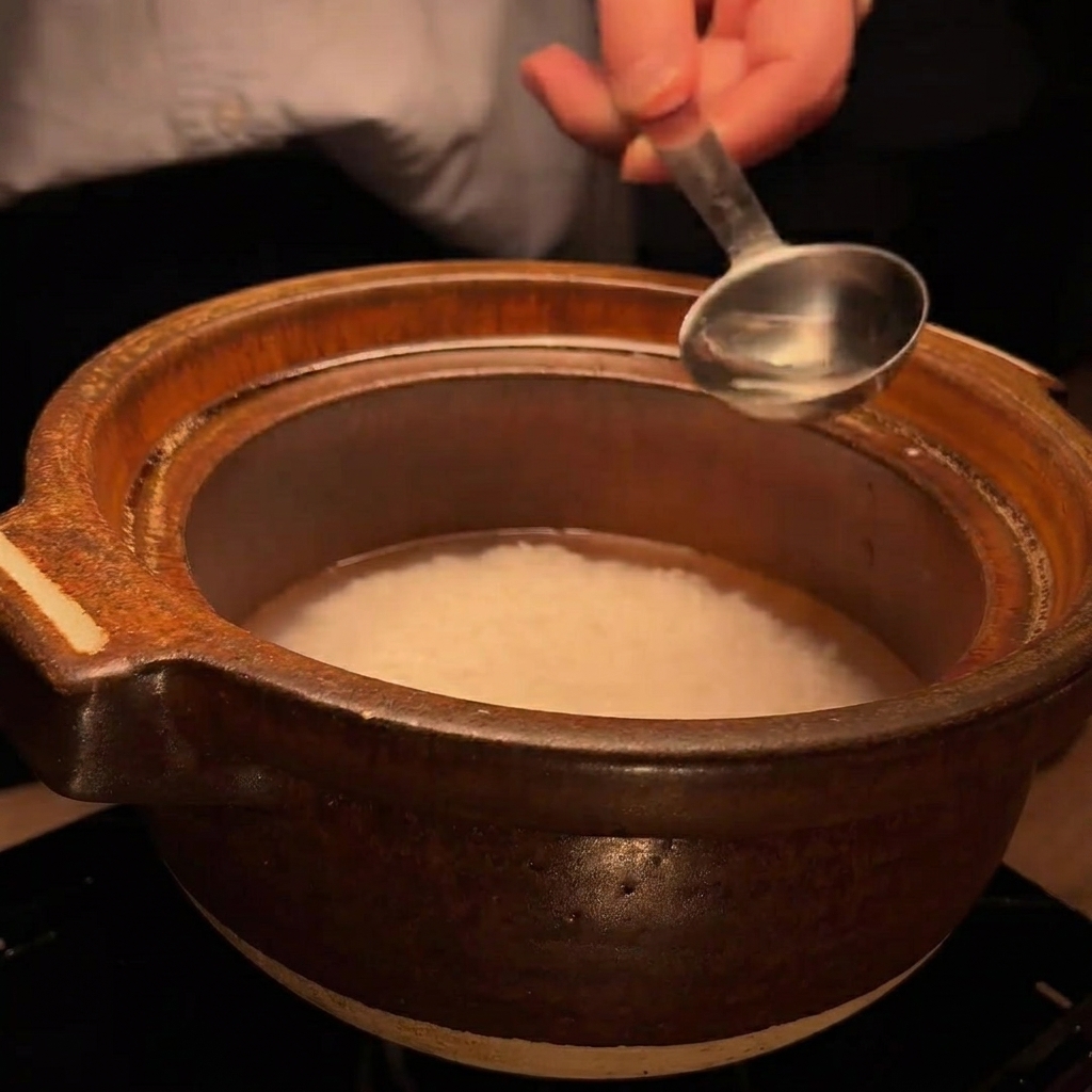 Une cuillère en métal verse une petite quantité de liquide clair, du saké, dans le pot en terre cuite avec le riz.
