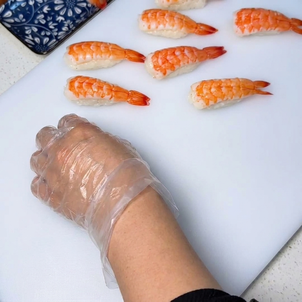 Gros plan dune crevette cuite placée sur une petite boulette de riz, pressée par des mains gantées pour former un sushi net.