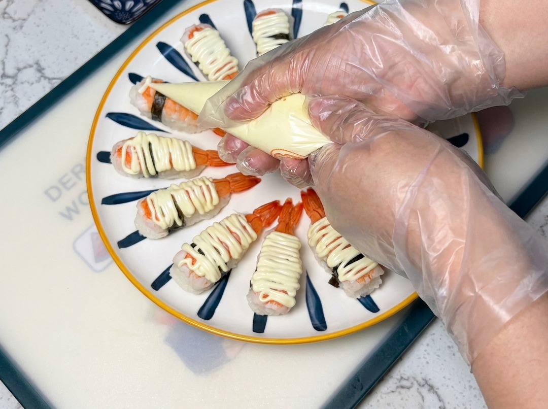 Gros plan sur une poche à douille déposant de la mayonnaise sur des sushis aux crevettes disposés sur une assiette.