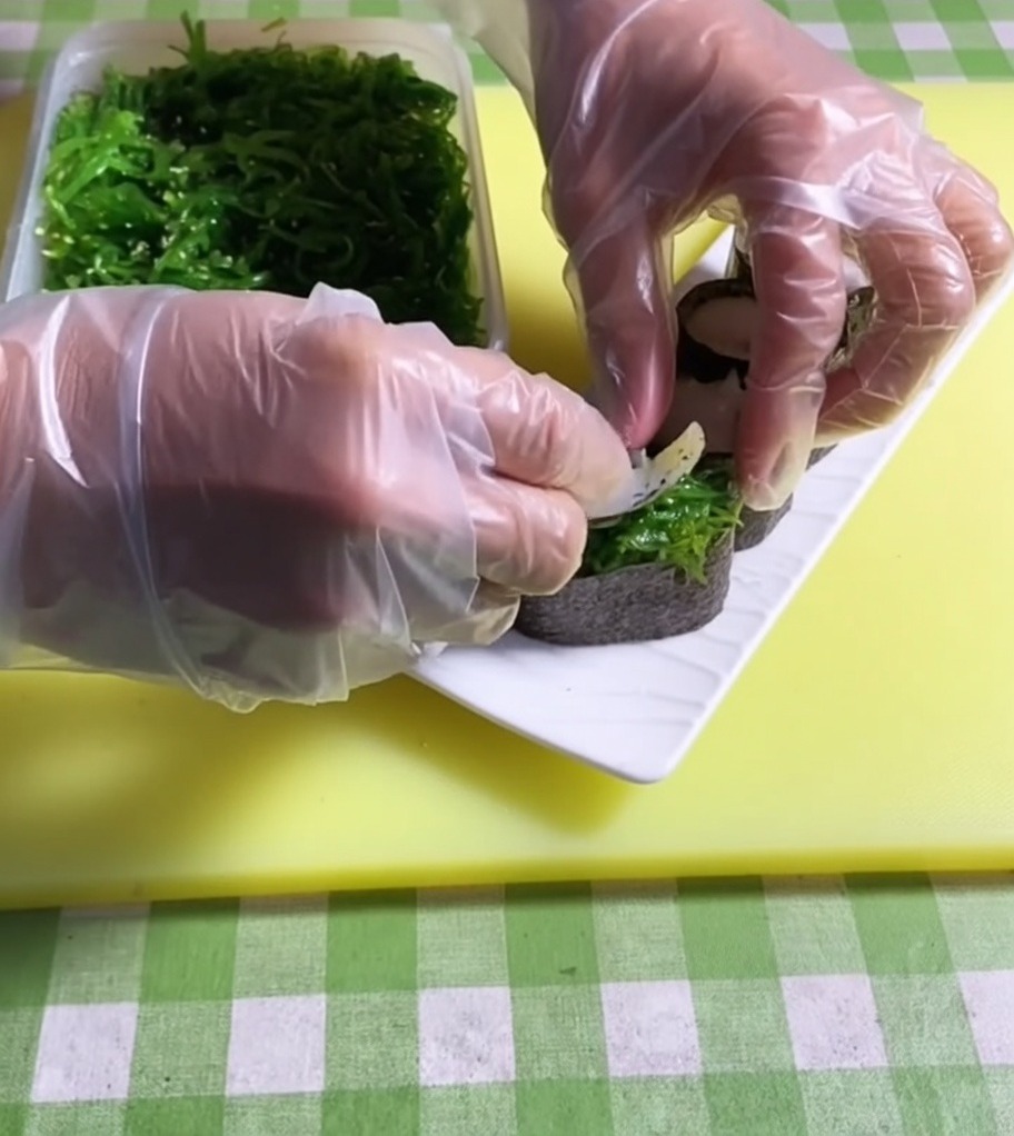 Une pince déposant une portion de salade dalgues vert vif sur le dessus dune base de sushi.