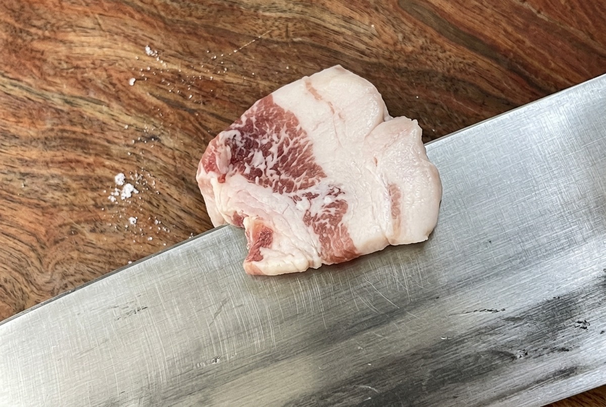 Un couteau bien aiguisé coupant un morceau de poitrine de porc crue en lamelles fines et uniformes sur une planche à découper en bois.