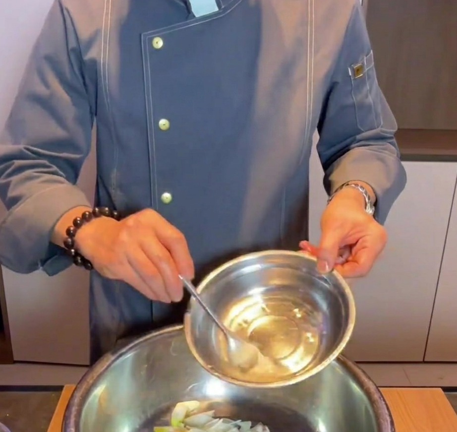 Une main versant de leau au bicarbonate de soude dans un bol mélangeur contenant de la poitrine de porc, du gingembre et de la ciboule.