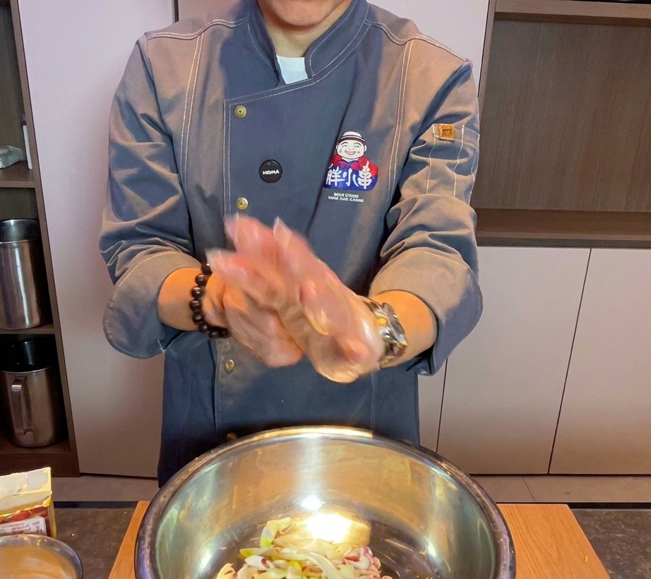 Un chef portant un gant transparent mélange la poitrine de porc et les aromates dans un grand bol en métal.