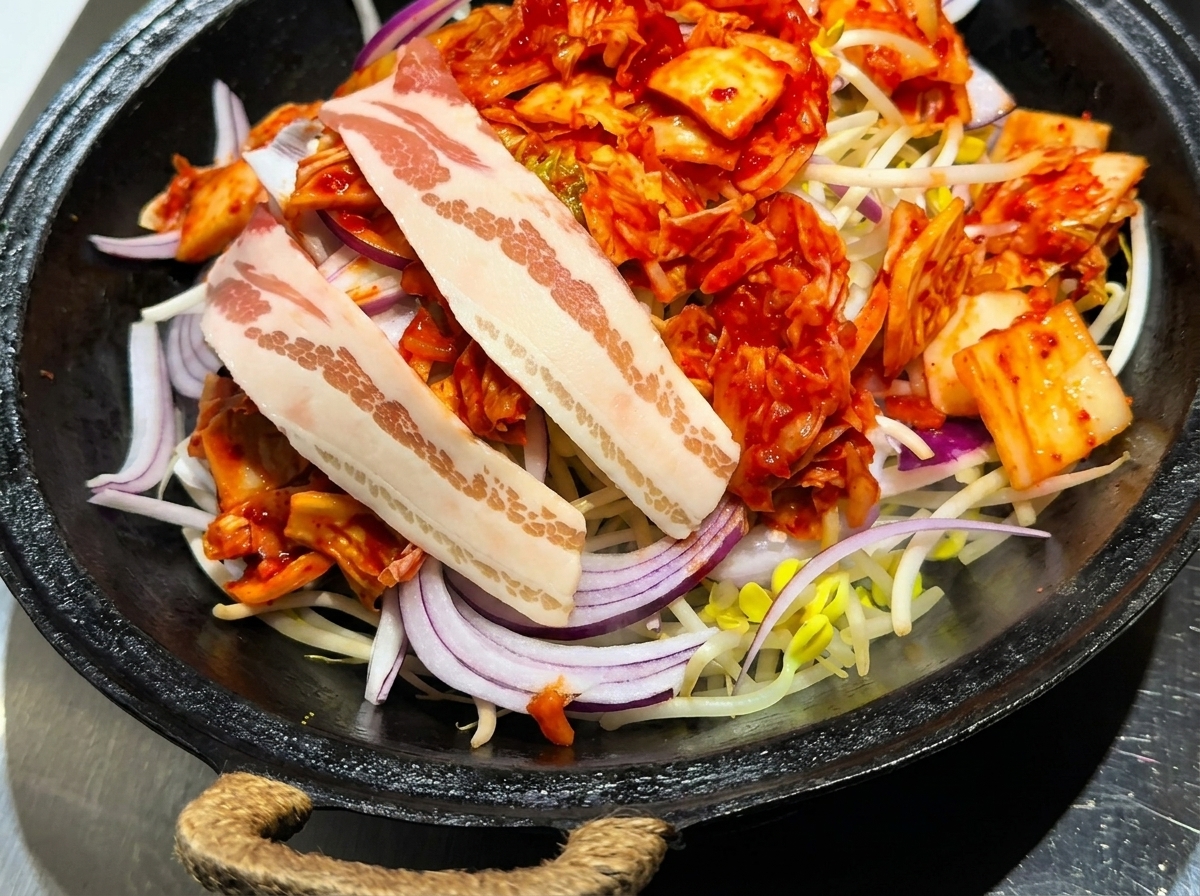 Tranches de poitrine de porc crue soigneusement disposées sur un lit de kimchi, doignons et de pousses de soja.