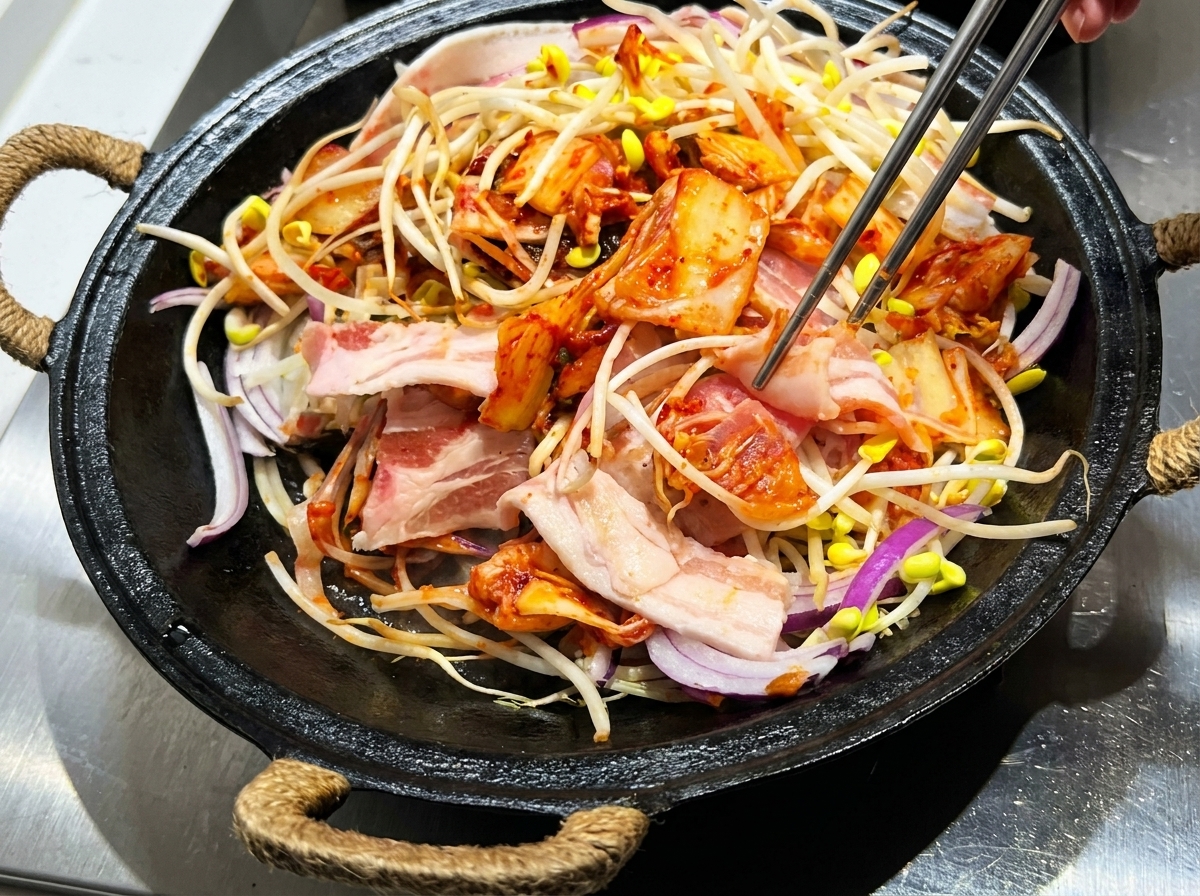 Pinces mélangeant la poitrine de porc, les pousses de soja, le kimchi et la sauce dans une cocotte en fonte.